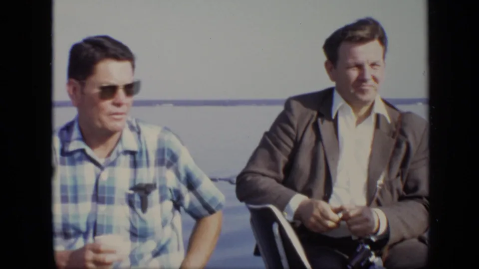 In the 1971 Moscow boat scene, the man in a suit jacket sits beside a man in sunglasses and appears to handle a camera.