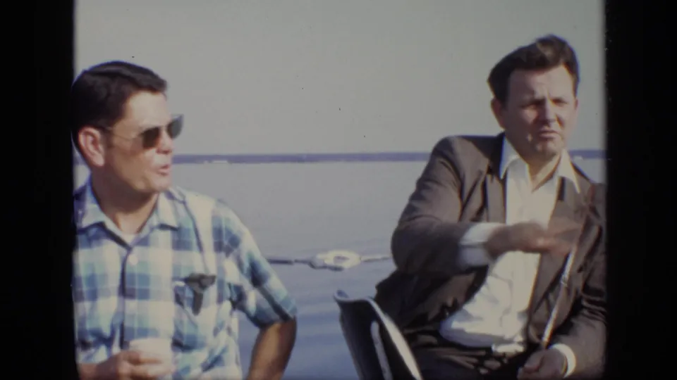 Two adult men remain seated on a boat in the Moscow home movie, with the suited man gesturing toward the water.