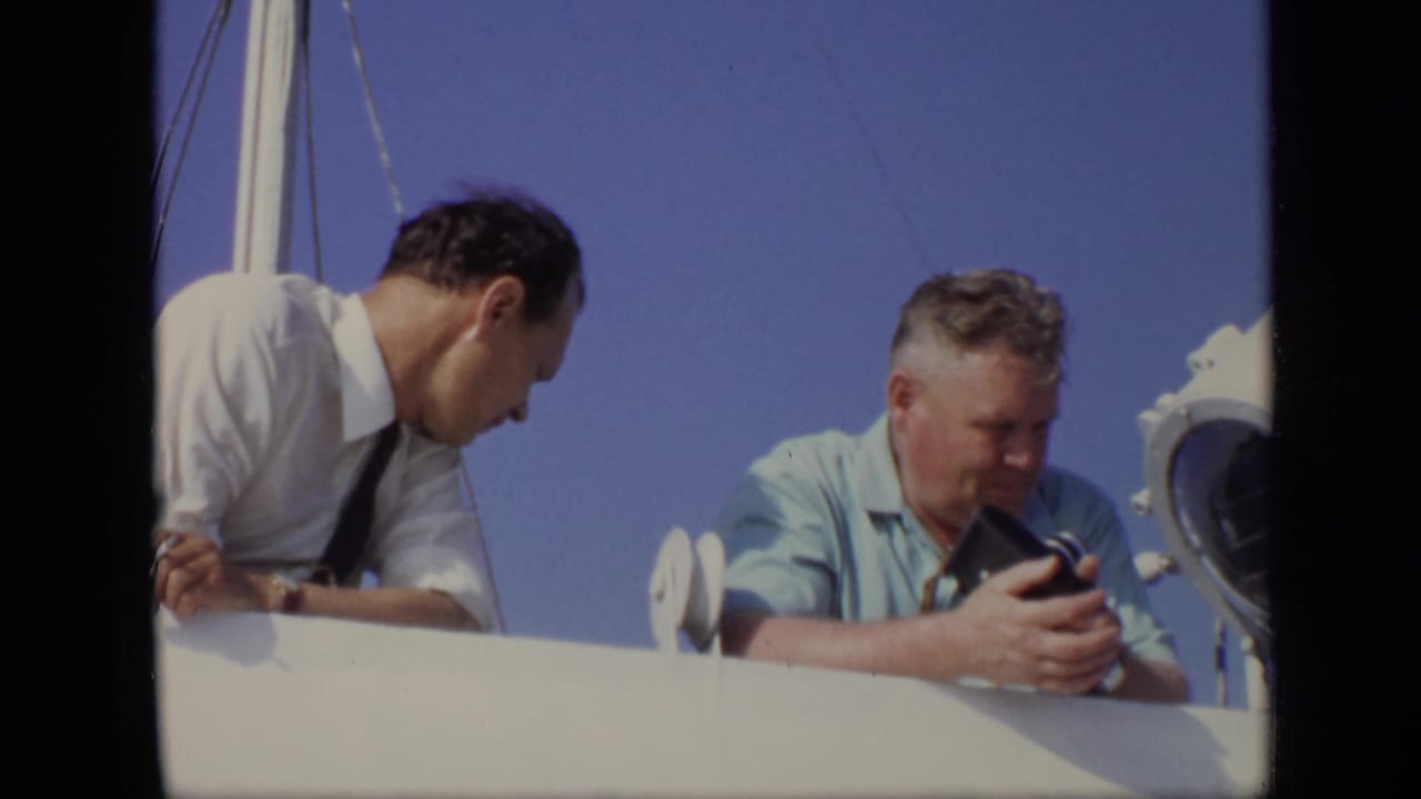 Two adult men lean over a white boat rail under a clear blue sky.