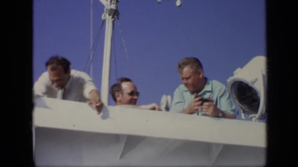 1971 8mm Moscow home-movie frame showing three adult men on a boat, with one in sunglasses between two men at the rail.