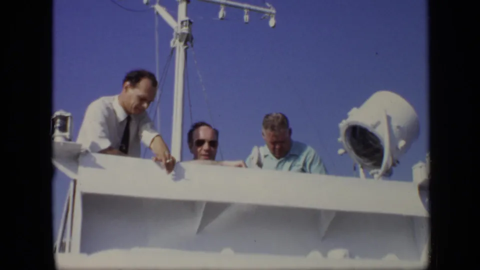1971 8mm Moscow frame of men aboard a boat, with a white rail, mast rigging, blue sky, and large spotlight visible.
