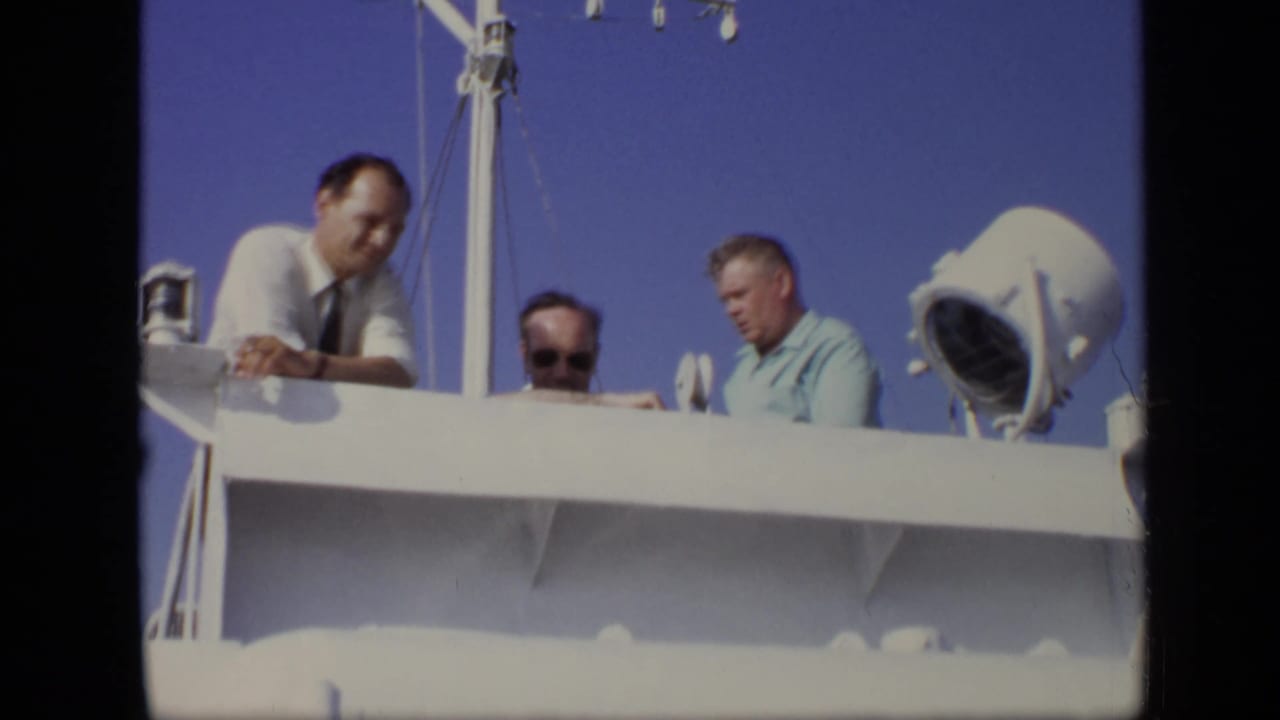 Three men viewed from a boat deck beside a white railing and deck light.