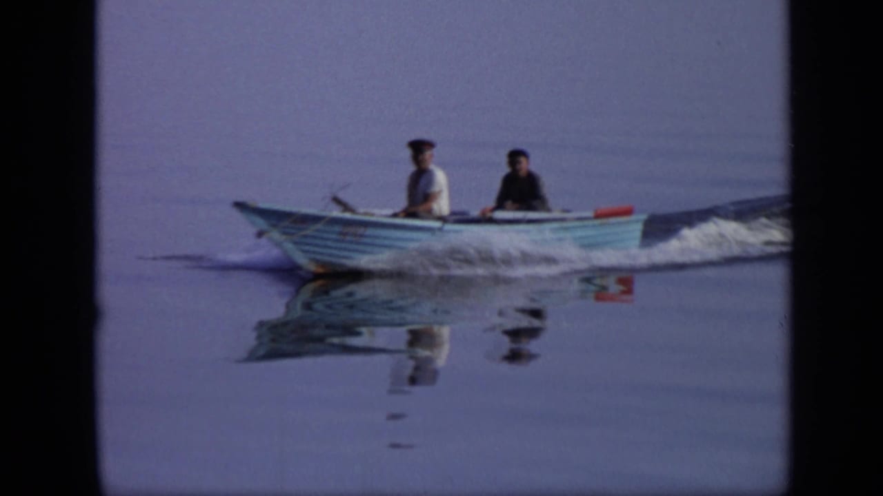 1971 Moscow home-movie frame showing two men in a small blue motorboat moving over still water with clear reflections below.