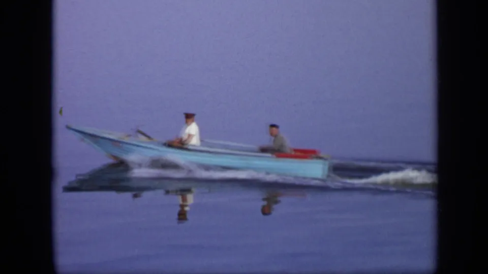 1971 Moscow home-movie frame showing two men in a blue motorboat moving left to right with a faint wake and water reflections.