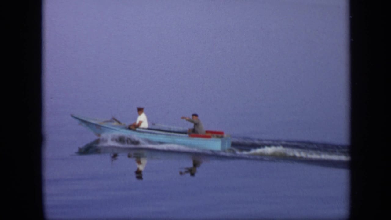 Two men ride in a small blue motorboat across calm water.