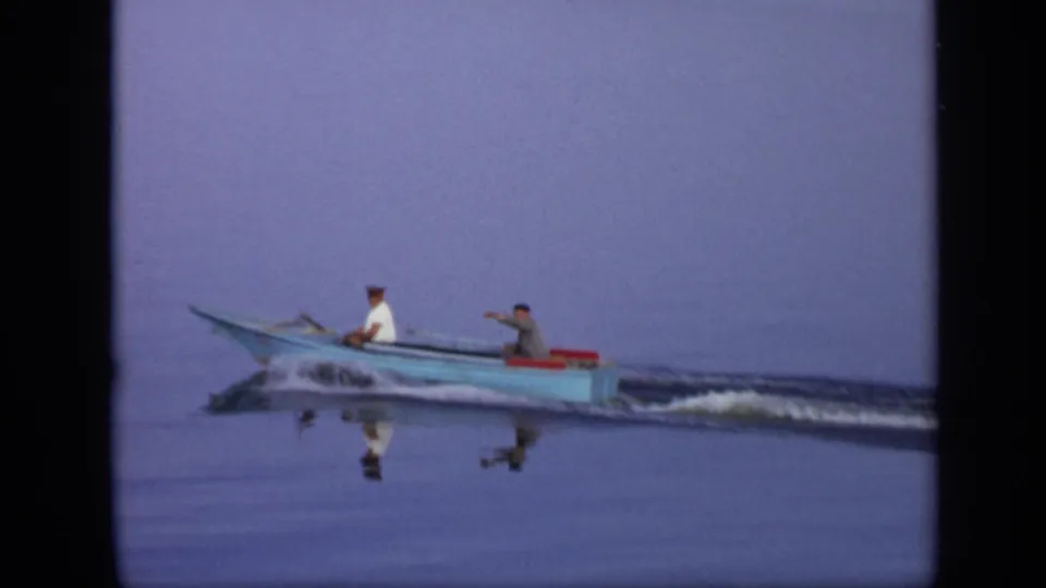 In supported 1971 Moscow home-movie footage, two men sit in a small blue motorboat moving across still water.