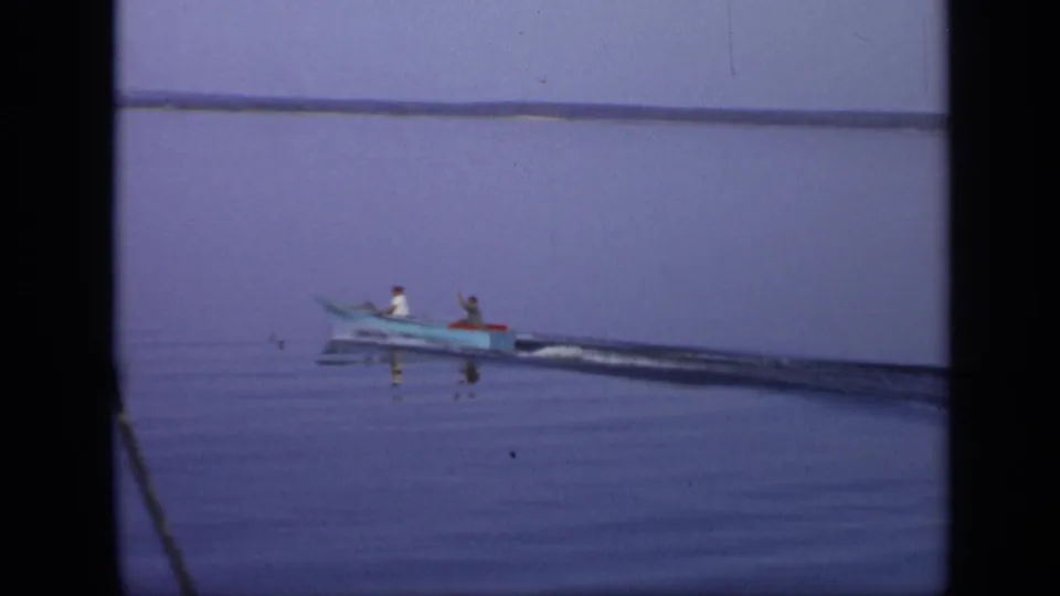 The small boat recedes across calm water in the 1971 Moscow footage, leaving a dark wake behind it.