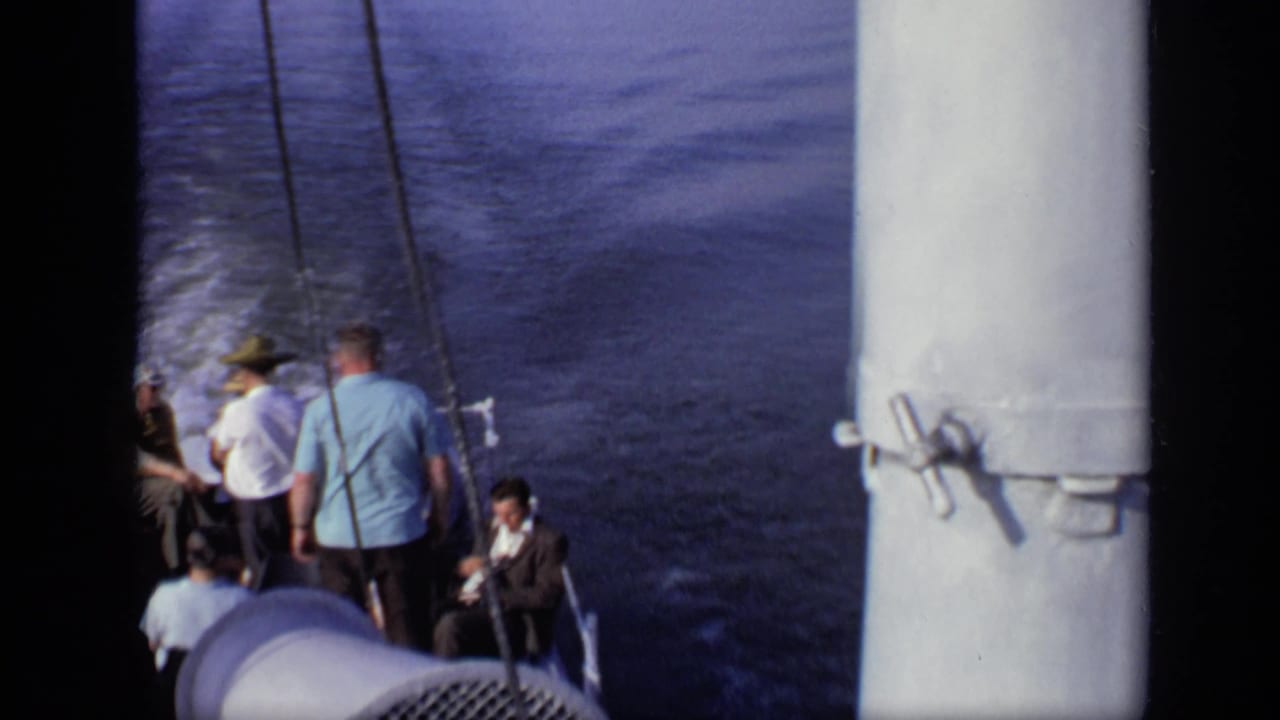 Passengers stand and sit near the stern of a moving boat with wake behind them.
