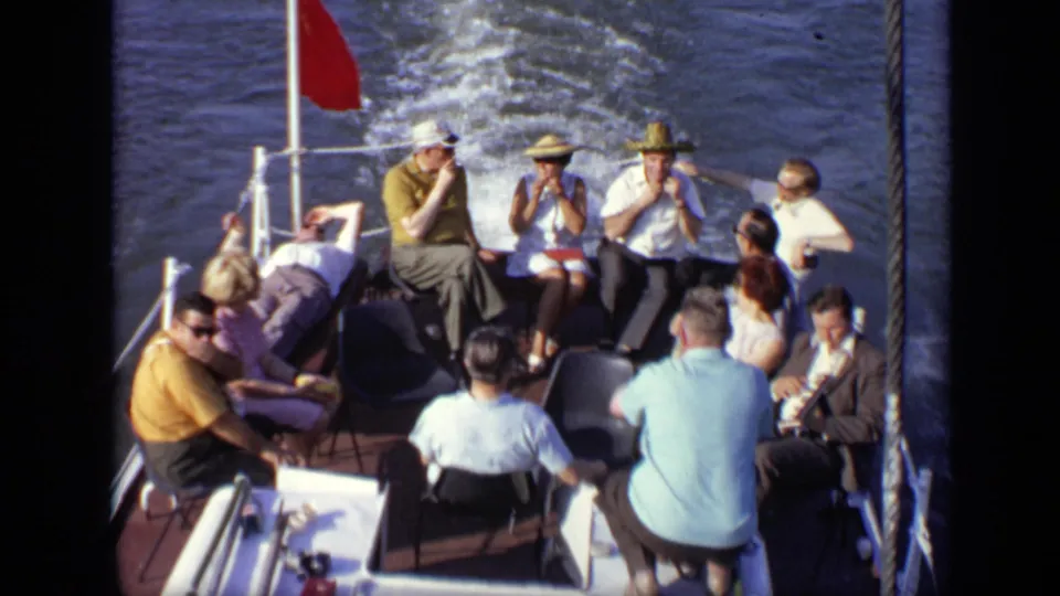 1971 Moscow home-movie frame showing adults seated on a boat deck, with a red stern flag and wake visible behind.
