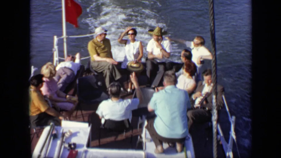 1971 8mm frame of adults on a Moscow boat ride, with one passenger adjusting sunglasses or shading their face near the stern.