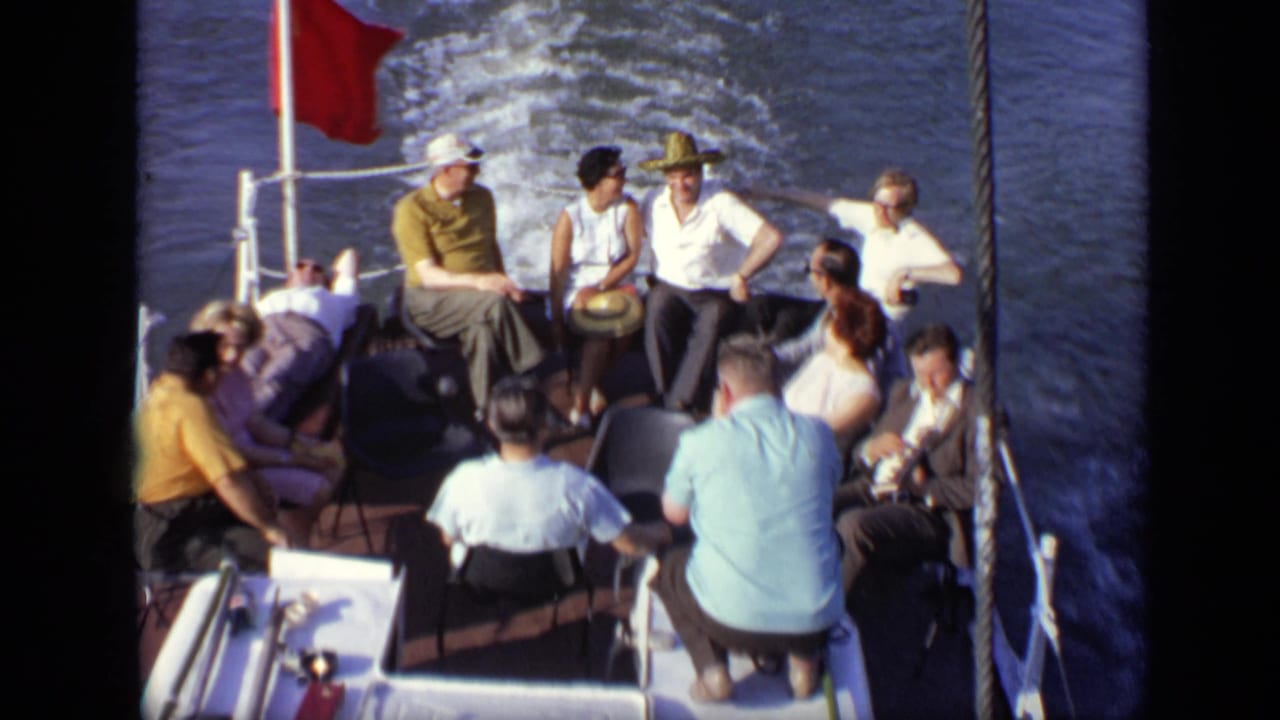 Group of adults on a moving boat, with water wake, rope rails, and a red flag visible.