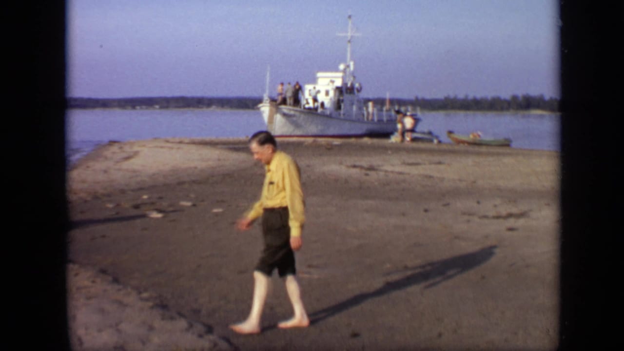 1971 Moscow-area 8mm frame of a barefoot man walking along a sandy riverbank near a white docked boat.