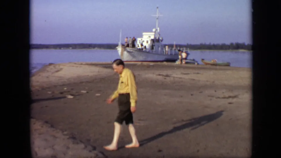 1971 Moscow-area 8mm frame of a barefoot man walking along a sandy riverbank near a white docked boat.