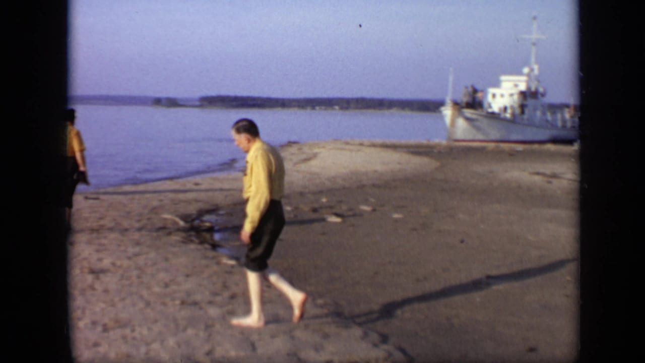 1971 Moscow home-movie frame showing a barefoot man on the riverbank with calm water and a white boat behind him.