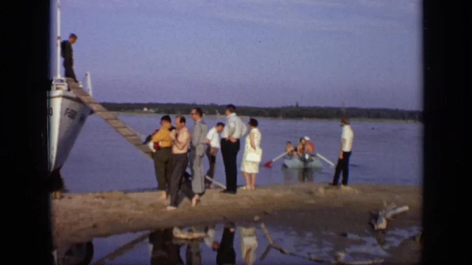 1971 Moscow 8mm frame of adults gathered at a river landing beside a docked white boat, gangplank, and small rowboat.