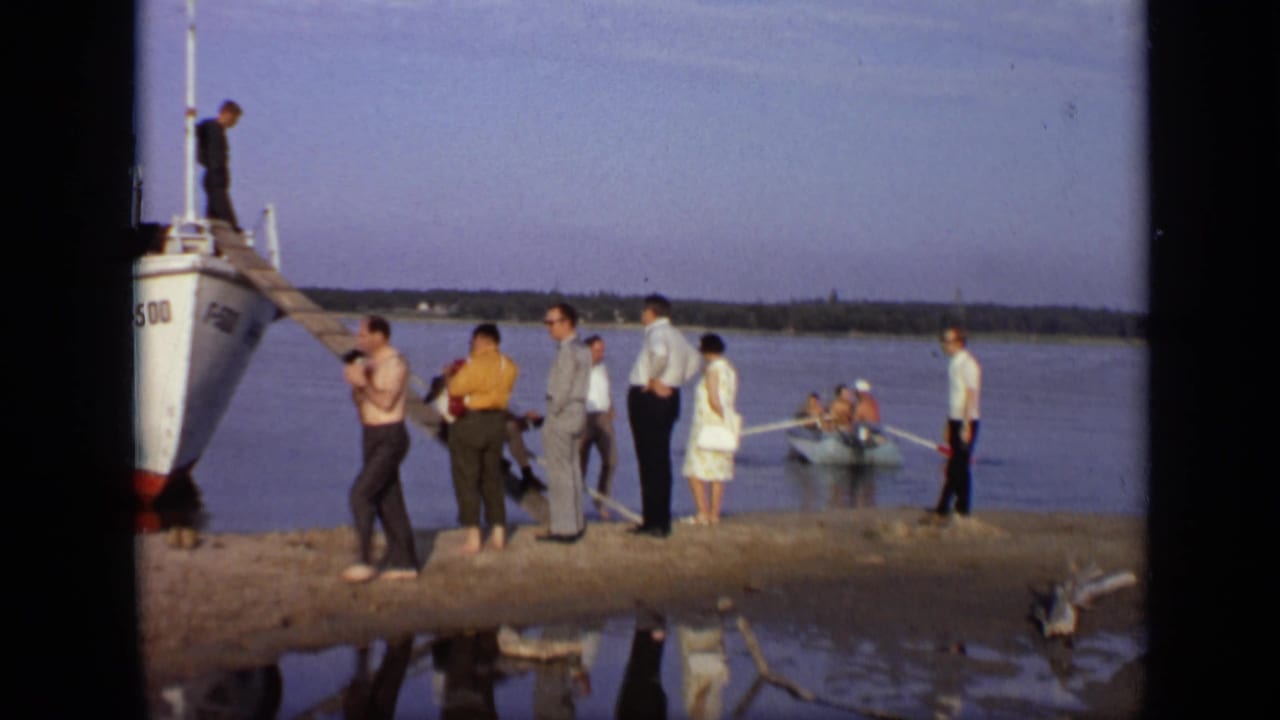People stand on a riverbank beside a docked white boat with a wooden ramp.
