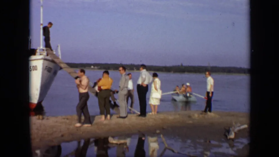 1971 Moscow home-movie frame showing people on a riverbank near a white boat, wooden gangway, calm water, and a small rowboat.