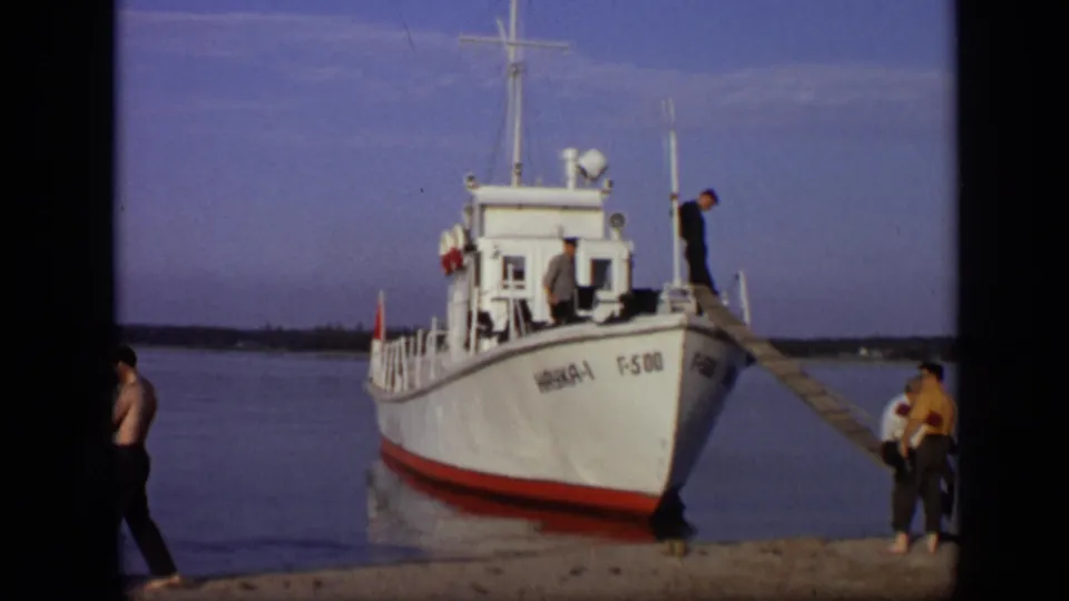 1971 Moscow home-movie frame with a white river boat at shore, a man on deck, a wooden gangway, and adults gathered beside the water.