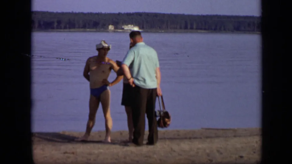 1971 Moscow lakeside frame showing two men watching the water from a sandy shore, with a distant boat visible.