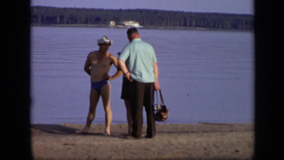 1971 Moscow home-movie frame of two men on a sandy lakeside shore looking toward calm water and a distant boat.