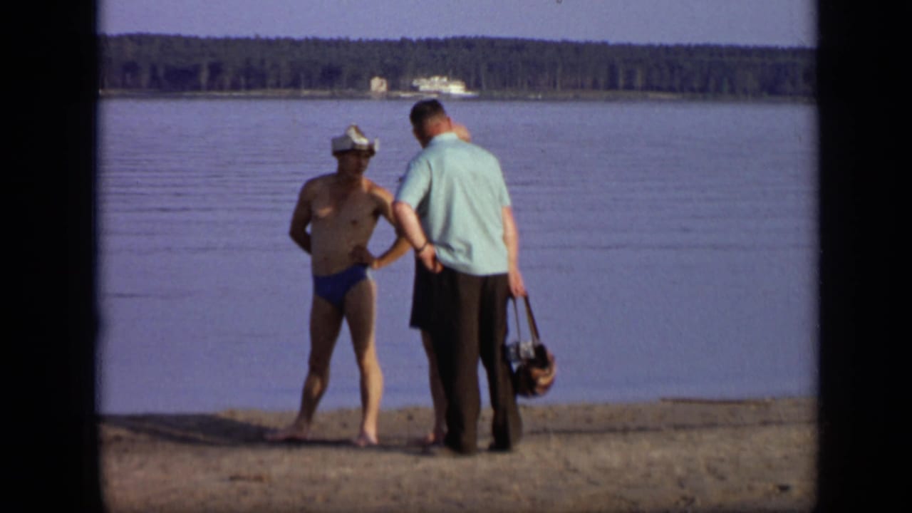 Two men stand near calm lake water on a sandy shore.