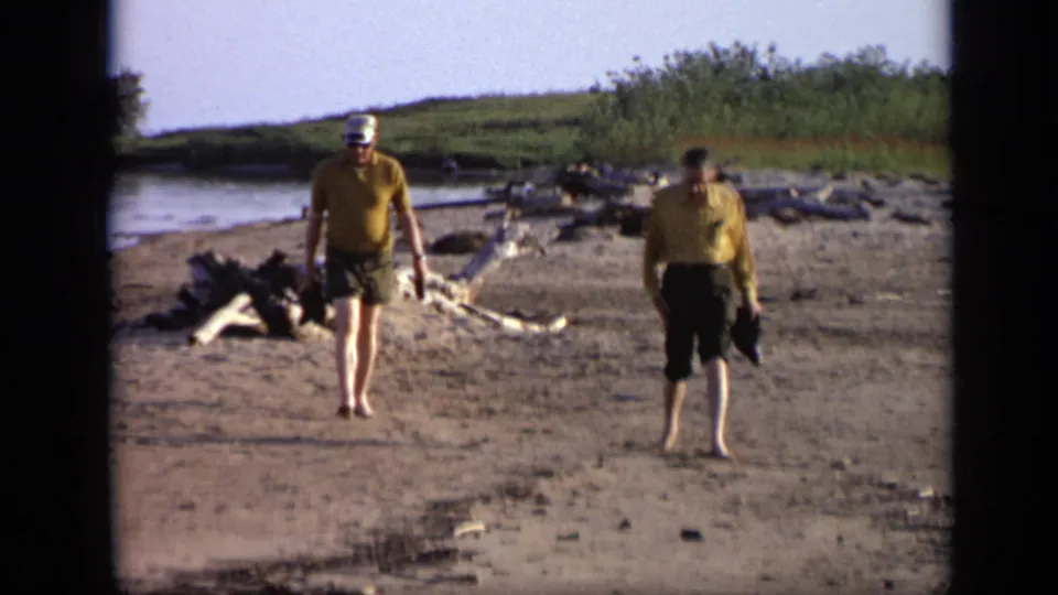 1971 Moscow home-movie frame with two men approaching the camera across a sandy lakeshore, with driftwood and brush behind them.