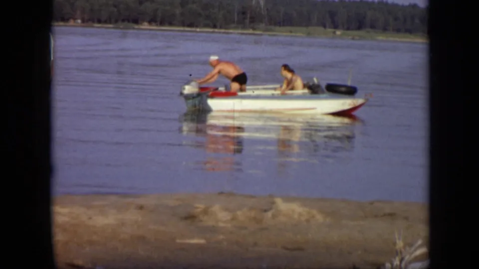 1971 Moscow lakeside frame showing two people in a small light-colored motorboat on still water near a sandy foreground.