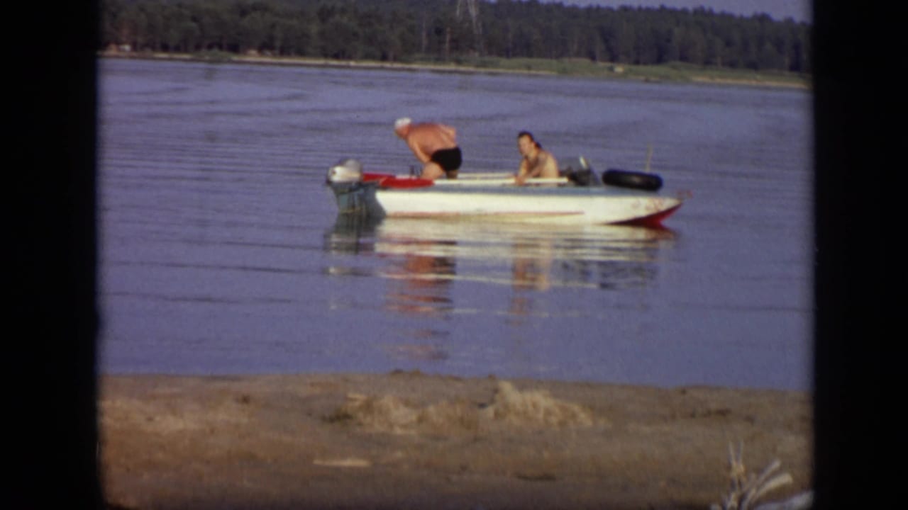 1971 Moscow home-movie frame showing two adult men in a white motorboat, one leaning near the outboard motor on a calm river.