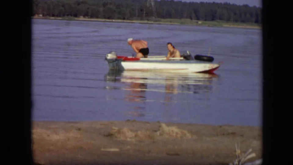 1971 Moscow home-movie frame showing two adult men in a white motorboat, one leaning near the outboard motor on a calm river.