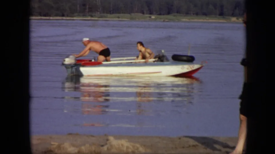 1971 Moscow home-movie frame showing a man in dark swim trunks leaning over the outboard motor as another man waits in the boat.