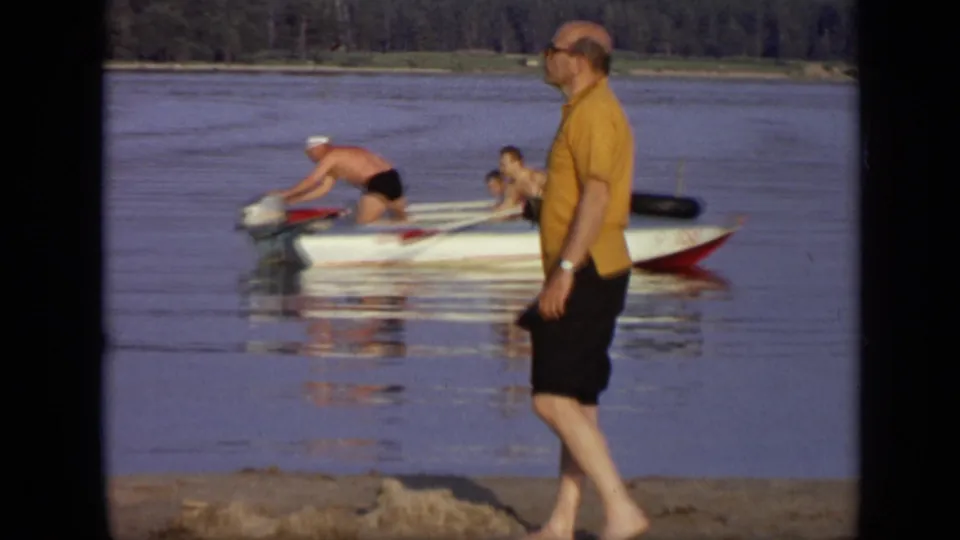 1971 Moscow frame with a man in a yellow shirt passing on the sandy shore while two men remain in the motorboat behind him.