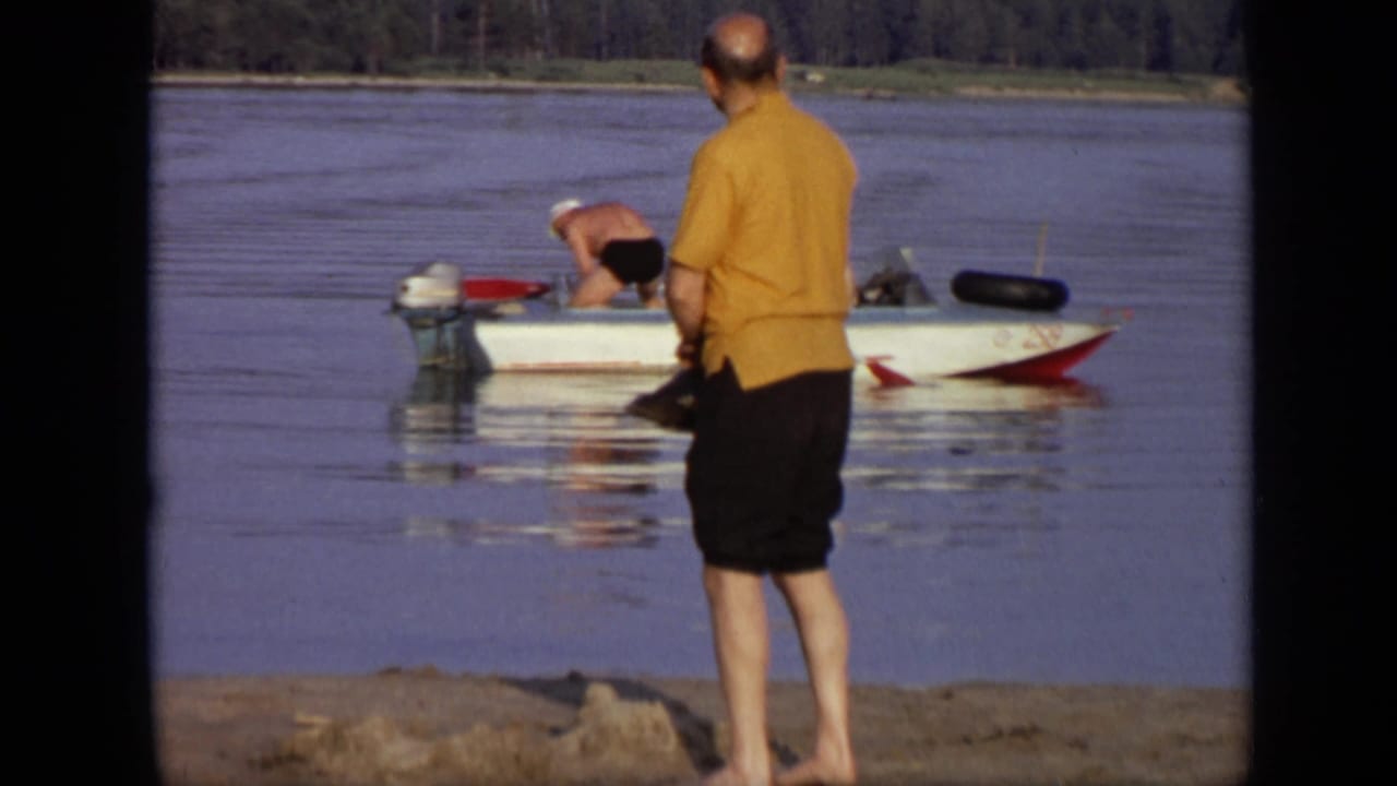 1971 Moscow home-movie frame showing a barefoot man in a yellow shirt near a small red and white motorboat on a calm waterfront.