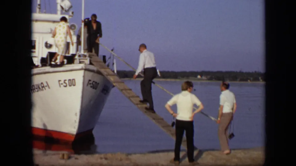 Passengers continue boarding the white river boat in the 1971 Moscow scene, while two men watch from the grassy bank.