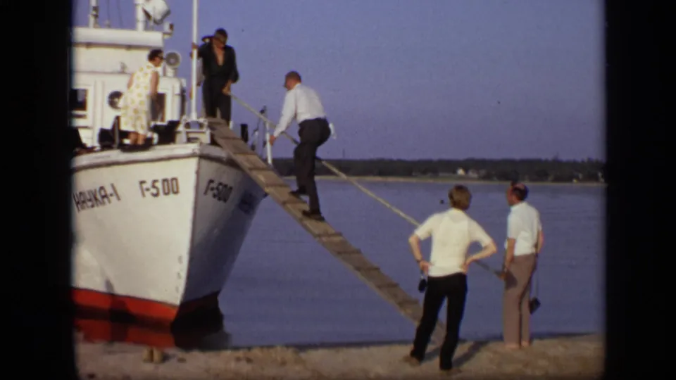 A man in a light shirt climbs the plank to the white boat as two adults wait on the Moscow riverside in 1971 home-movie footage.