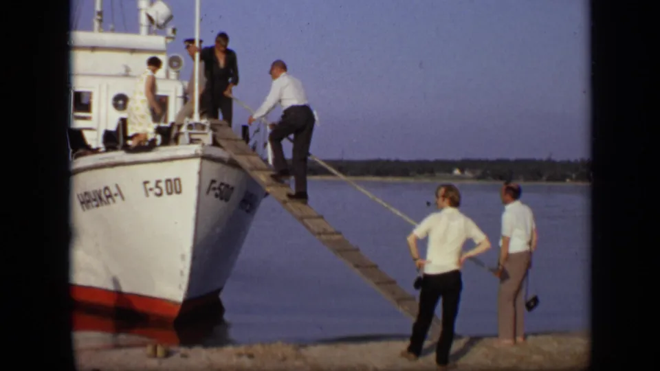 1971 Moscow home-movie frame showing adults near a white boat marked in Cyrillic as a man begins boarding by gangplank.