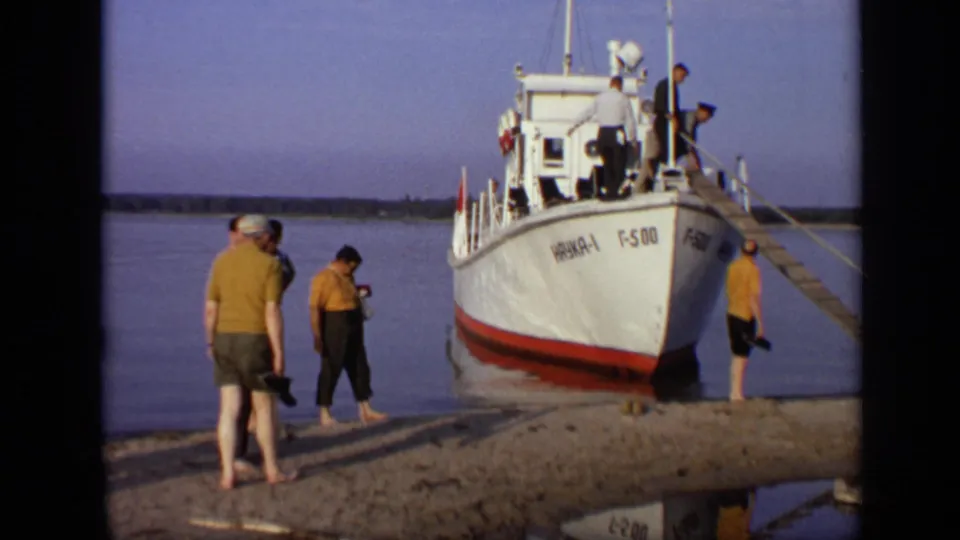 Wide 1971 Moscow home-movie view of adults around a moored white boat, with gangplank, calm water, and shoreline visible.