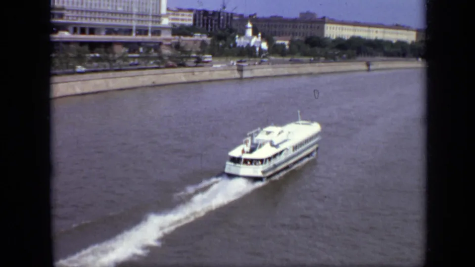 Passenger ferry travels along a broad Moscow river with waterfront buildings and a pale domed church form in the distance.