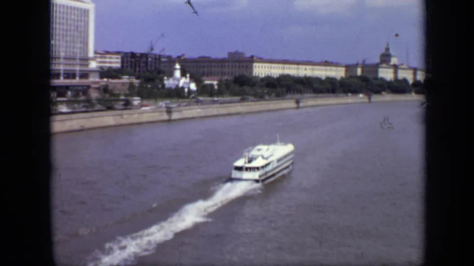 White passenger ferry recedes across the Moscow river, framed by embankment, city blocks, and a small domed church shape.