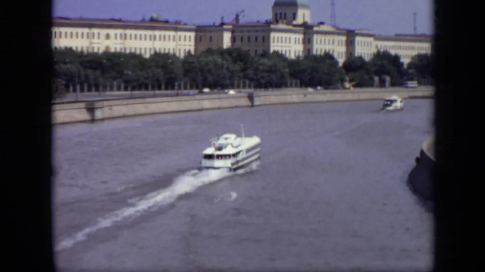 1971 Moscow home-movie frame with a ferry, river wake, embankment trees, and a domed roofline beyond.