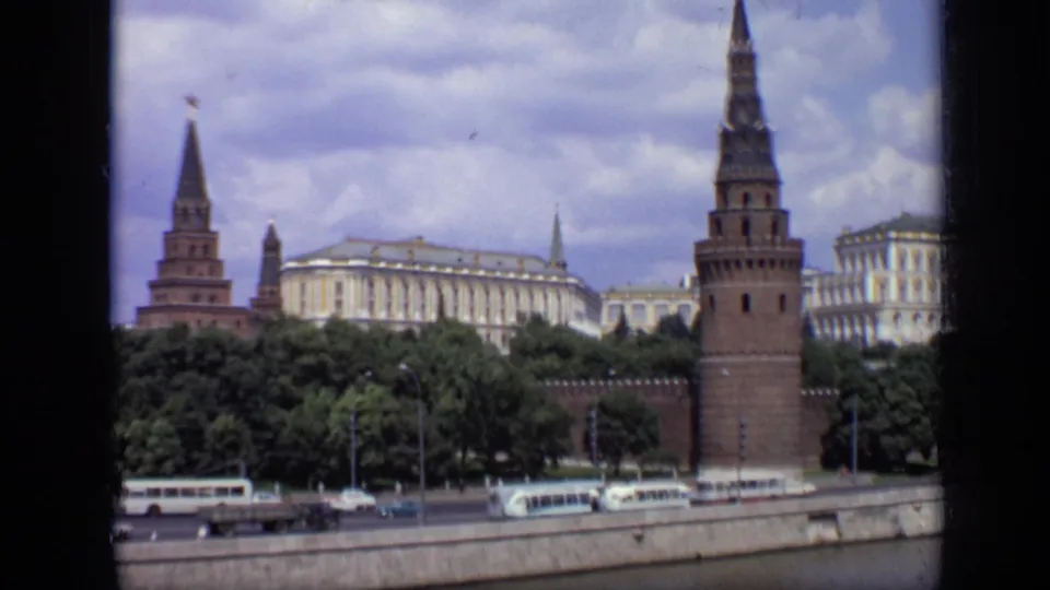 1971 Moscow home-movie frame showing Kremlin towers above a tree line and Soviet-era traffic along the river embankment.