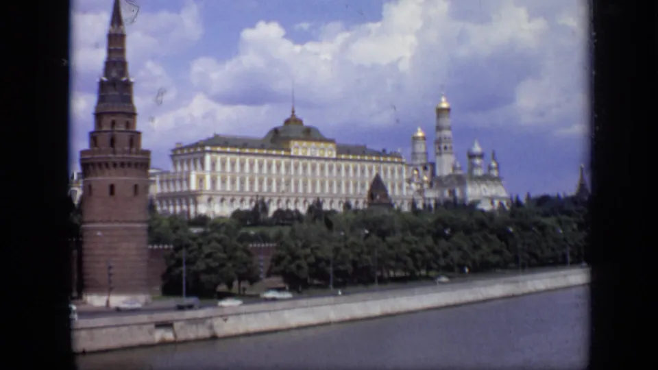 1971 Moscow 8mm frame showing a wide Kremlin view with tower, white facade, green trees, road, and river edge.