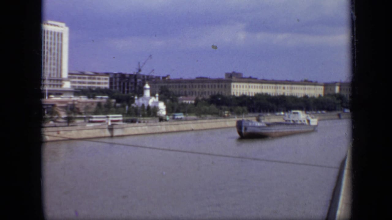 Wide river view with a barge approaching a Moscow embankment, buildings, cranes, and a white domed church.