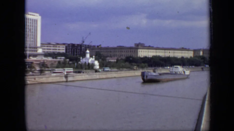 1971 Moscow riverfront seen from an elevated moving viewpoint, with a barge on the river and Soviet-era buildings behind the embankment.
