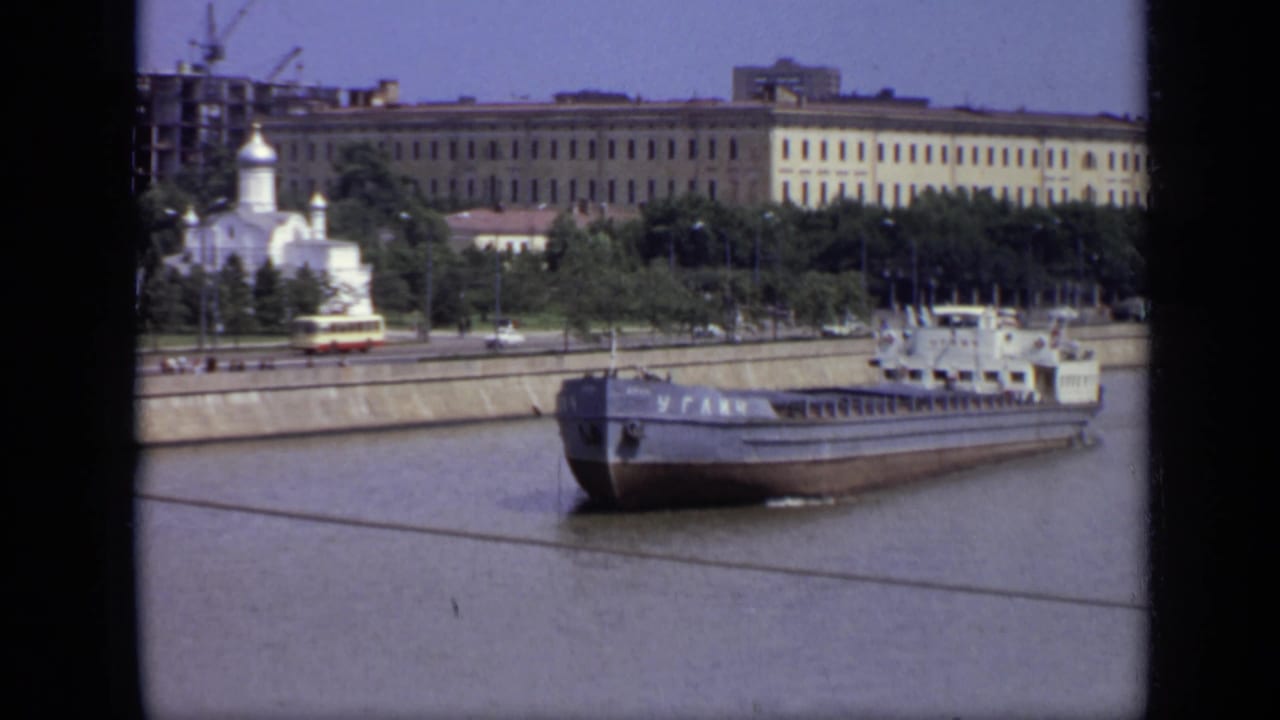 Closer river view of a barge passing an embankment with trees, road traffic, buildings, and a white church.