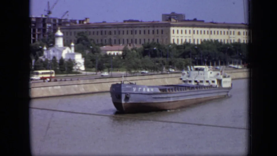 The barge continues past the Moscow riverbank in 1971, framed by home-movie gate edges and background civic buildings near a white domed church.