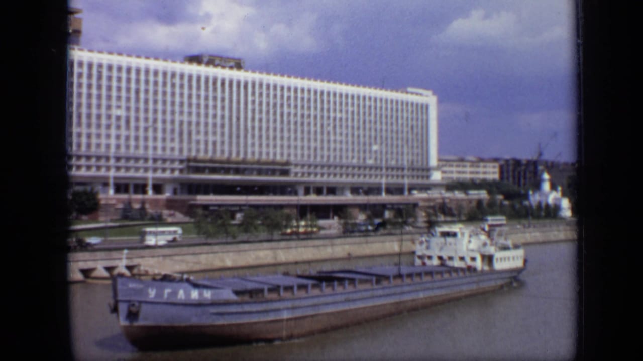 Barge on a Moscow river with buses and a large windowed building behind it.