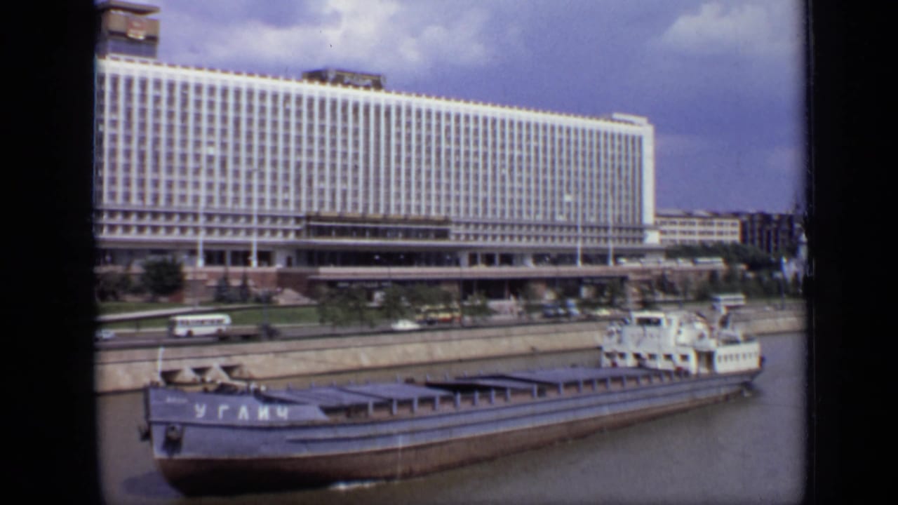 1971 Moscow view of a barge marked in Cyrillic passing an embankment road with buses, cars, and a broad modern building.