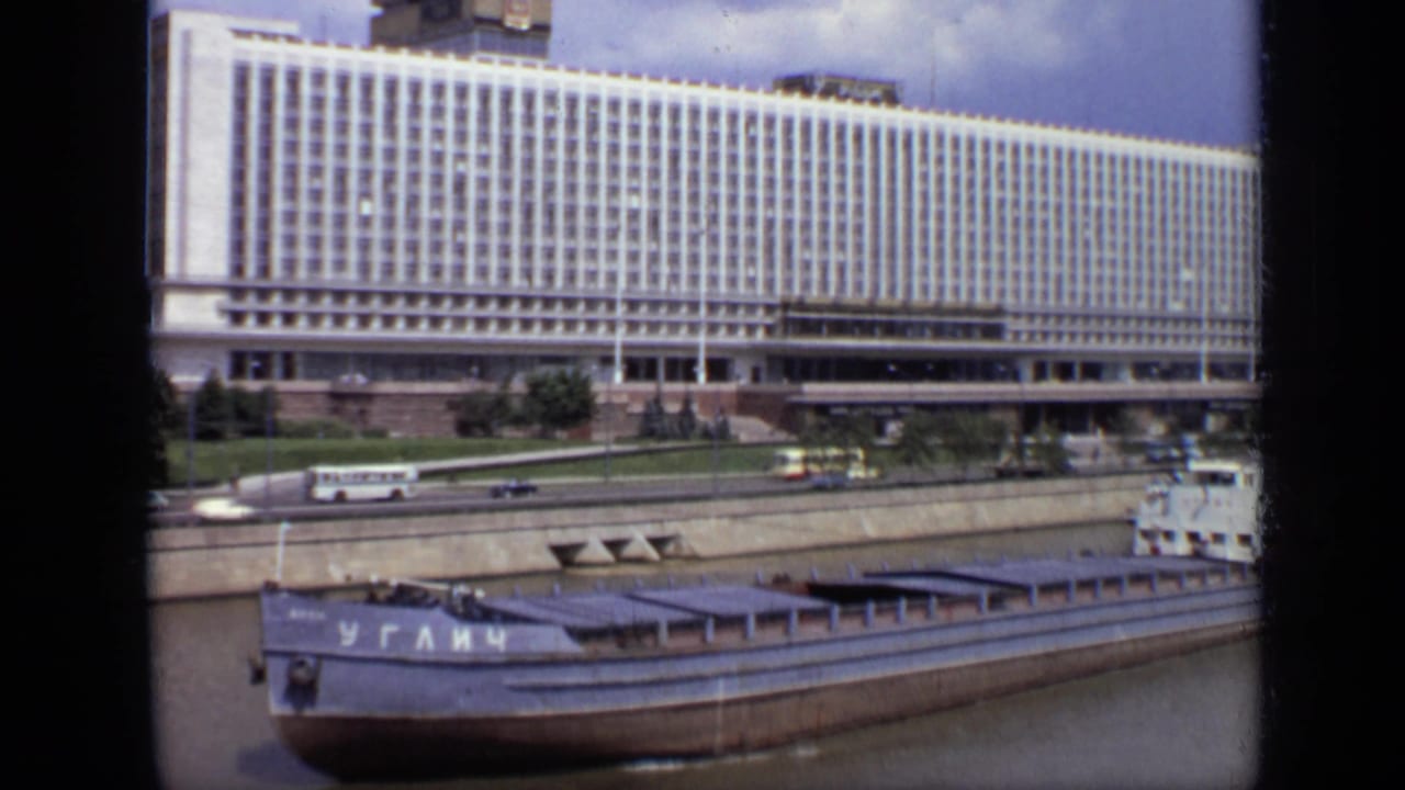 Cyrillic-marked barge on a river with a large white building and traffic behind it.