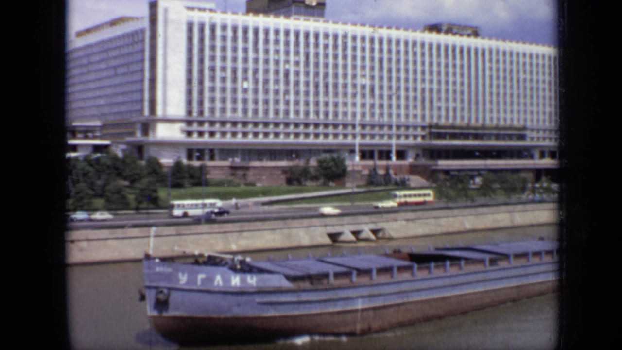 Wide riverside view with barge, embankment traffic, trees, and a large gridded building.