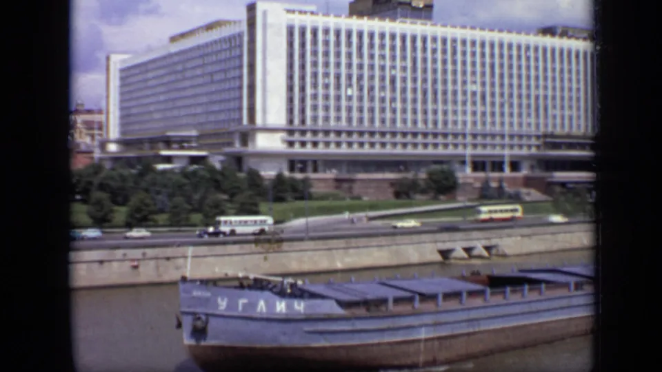 1971 Moscow 8mm view of a river barge moving past an embankment road, with small pedestrians, buses, cars, trees, and a large white building.